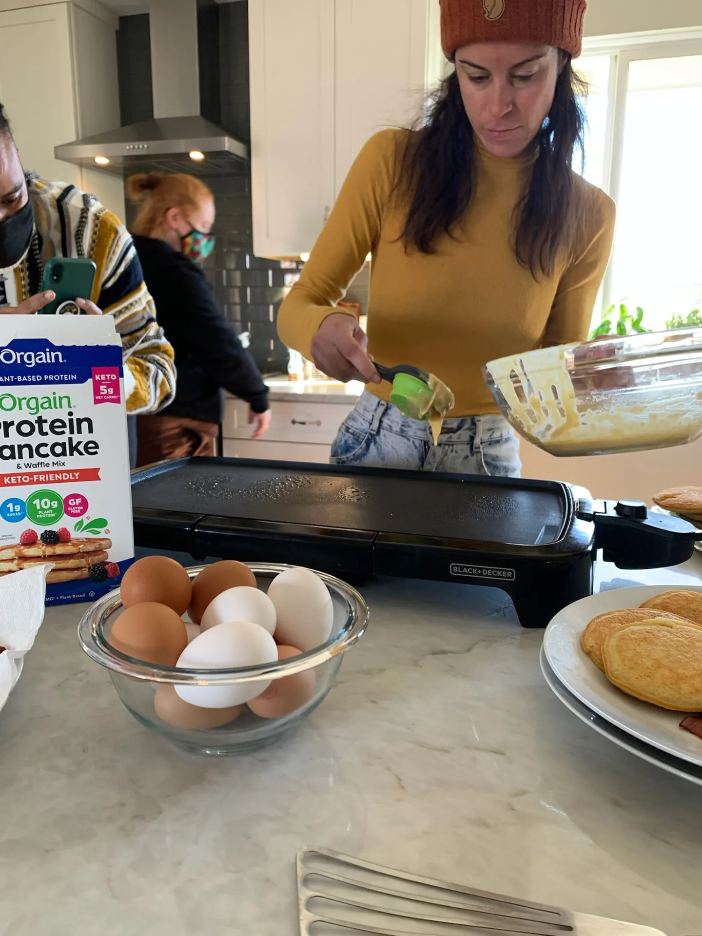 Orgain protein pancake mix on a kitchen counter beside a griddle and eggs